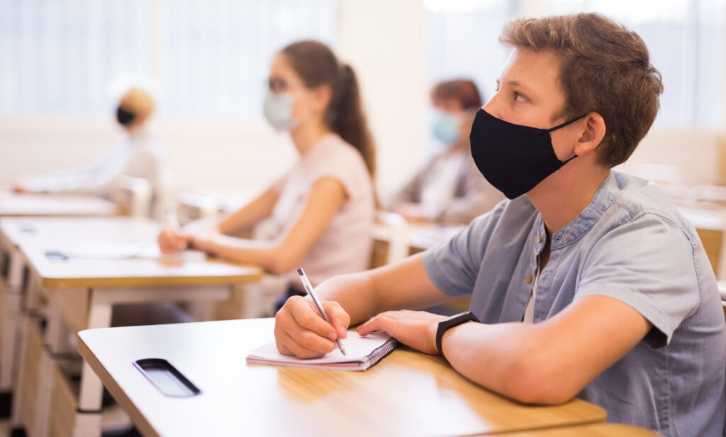 Teenager in protective mask writing in workbook on lesson