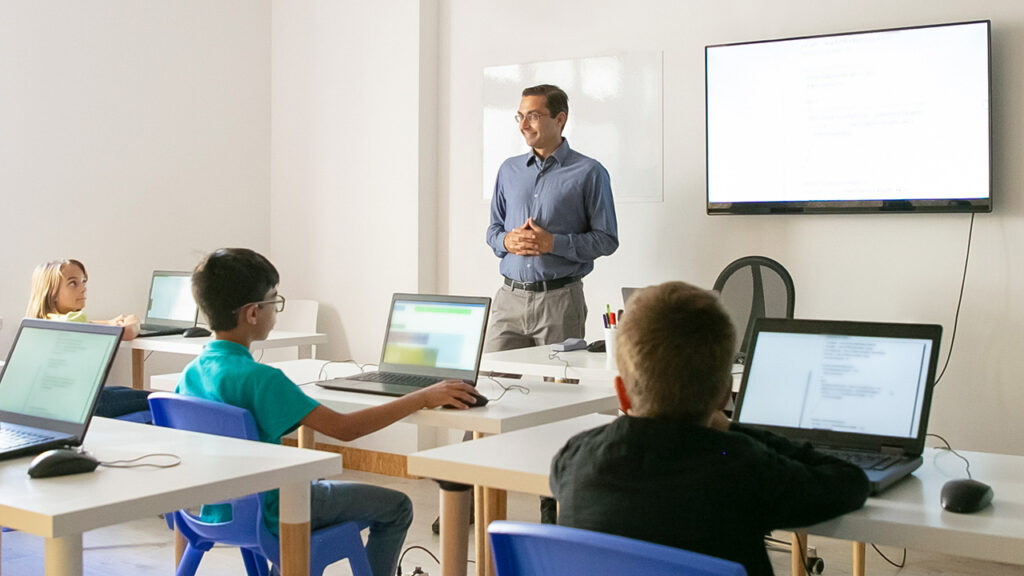 A teacher stands in front of a classroom filled with children looking at laptops