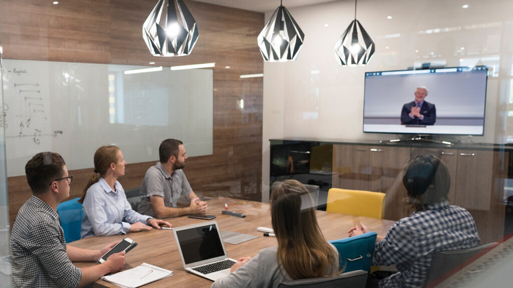 A group of employees in a conference room watch a man presenting on a television screen.