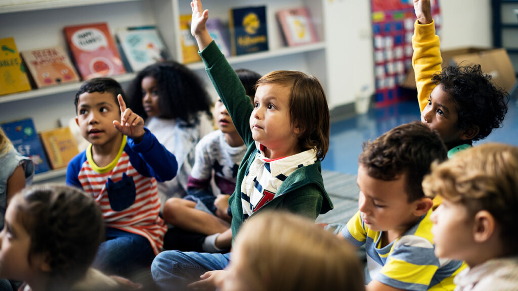 A classroom filled with young students sitting on the floor. A young girl raises her hand as if to ask a question.