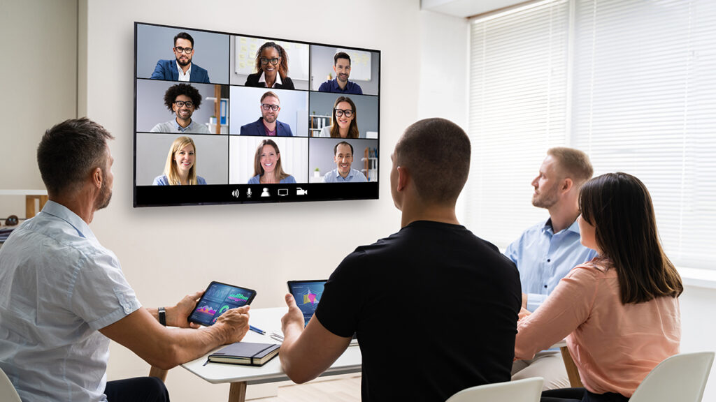 a group of people sitting in a conference room with a teleconference on the tv screen.